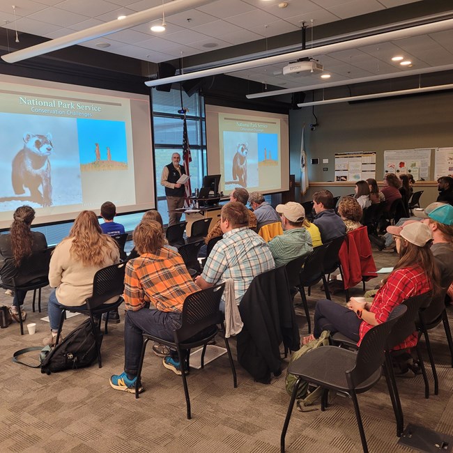 A sitting audience watches a speaker presenting at the front of the room