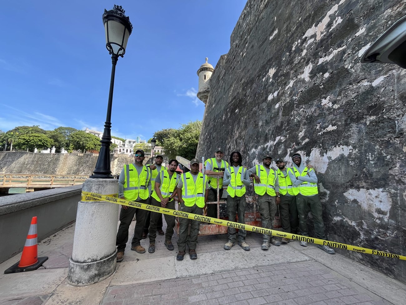 Nine workers stand behind caution tape tied to lamp post. Grey fort wall on the right. Workers wear green hats, green cargo pants, grey shirts, with neon green safety vests.