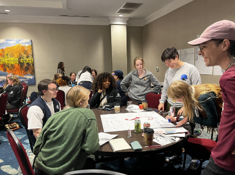 people sitting at a table smiling and working on a poster that is spread out in front of them, similar tables of others behind them.