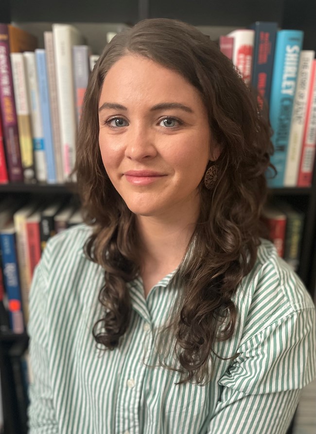 A white woman with long brown hair posing in front of a bookshelf