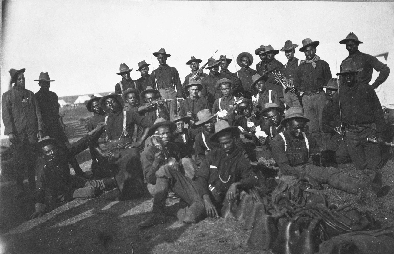 Black and white photo of African American soldiers in four lines posing for the camera. They are wearing military uniforms of 1898