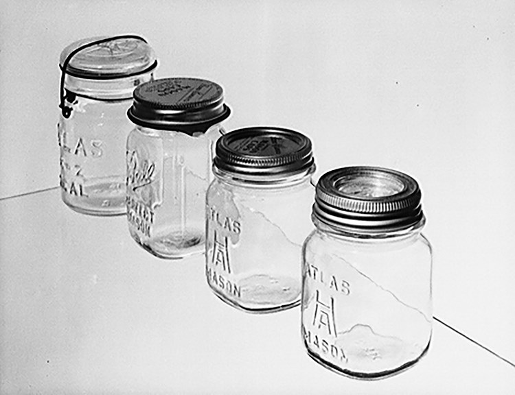 Black and white photo of four different types of canning jars, including one with a bail, one with a sealing ring, one with a metal cap and liner, and one with a metal ring and glass liner. Three are marked Atlas glass company; one is marked Ball.