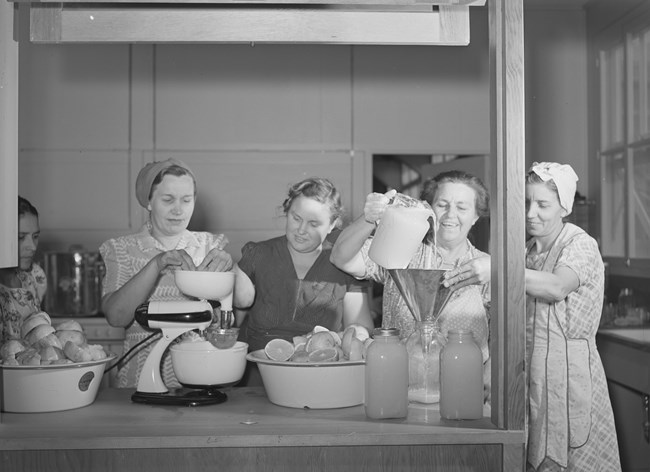 Black and white photo of five women working in a community kitchen. The woman on the far left is brown; the rest are white. One is juicing grapefruits, two are pouring juice into canning jars.