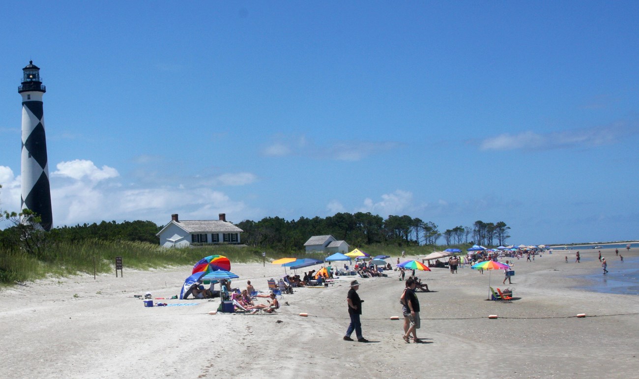 A sunny beach with a black and white diamond-patterned lighthouse on the left and many people under colorful umbrellas.