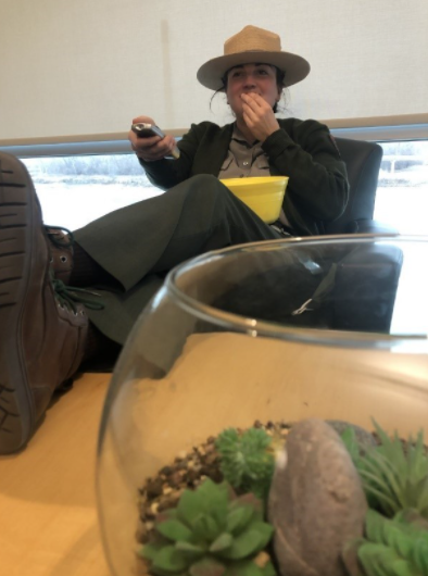 A ranger sits reclined in a chair. She is eating popcorn and holding a remote control. There is a terrarium with succulents in the foreground.