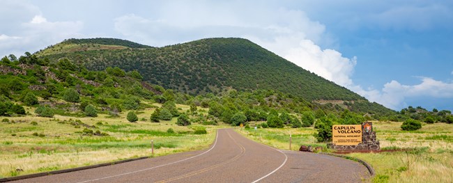 Entrance to Capulin Volcano National Monument with sign and cinder cone volcano covered with green vegetation under blue sky