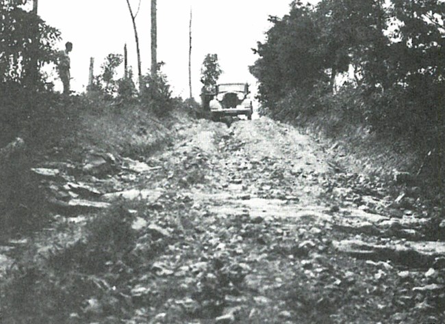 Black and white photograph of a car driving on a dirt road filled with deep ruts and large rocks.