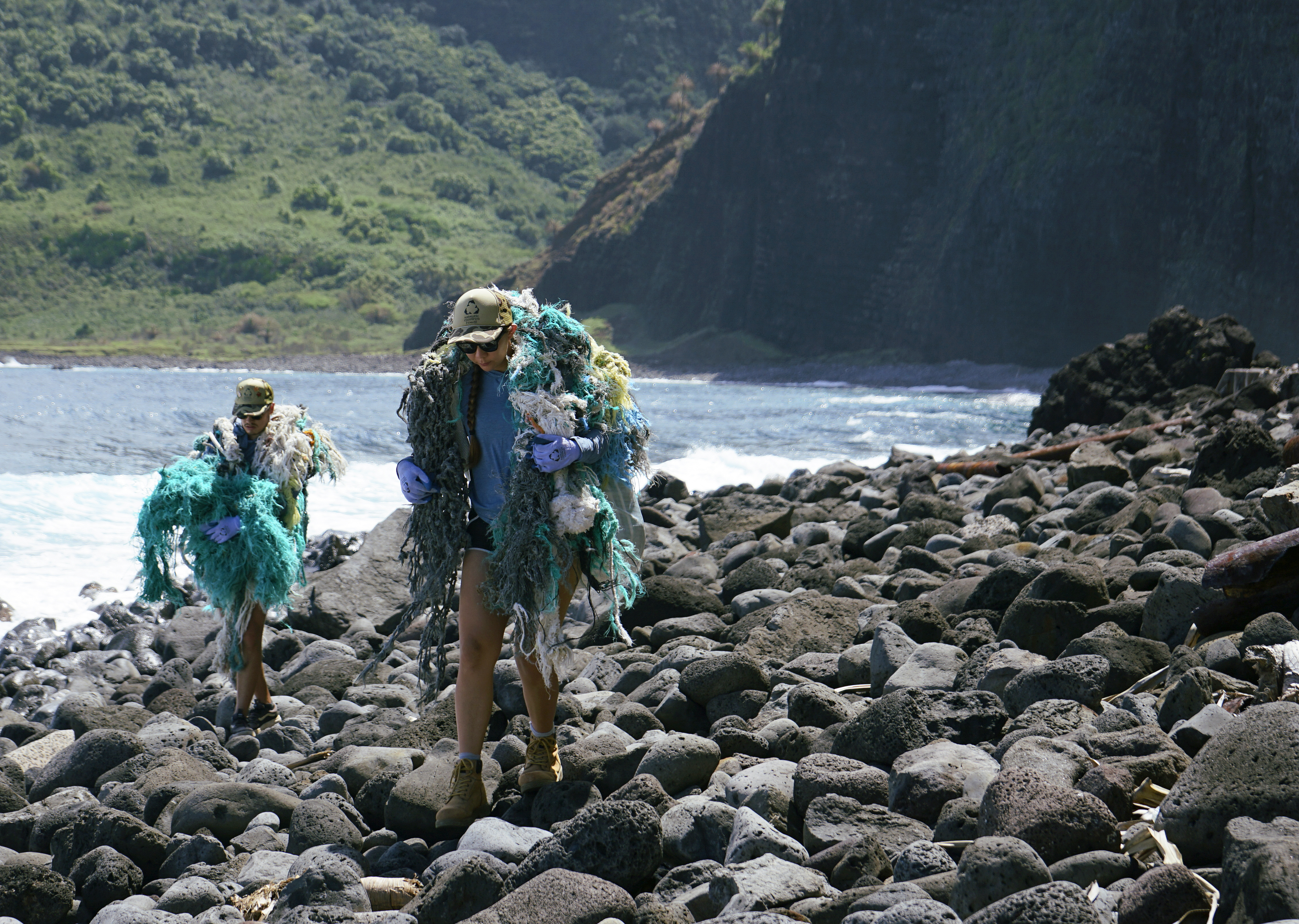 two people carry large bundles of collected marine fishing nets and other marine debris along a rocky shoreline with steep mountain cliffs in the background