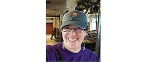 Cooper in an NPS ballcap and purple T-shirt, with mill equipment behind her.