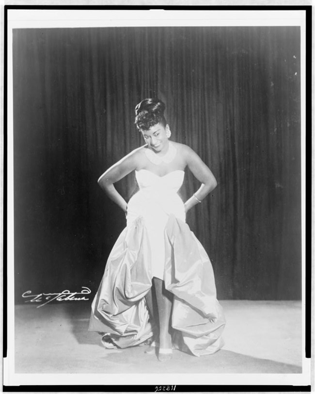 Full body black and white portrait of Celia Cruz on stage. Cruz wears a dress with a sweetheart neckline and high-low skirt. She rests her hands on her hips with her head tilted down, but still smiling up at the audience.