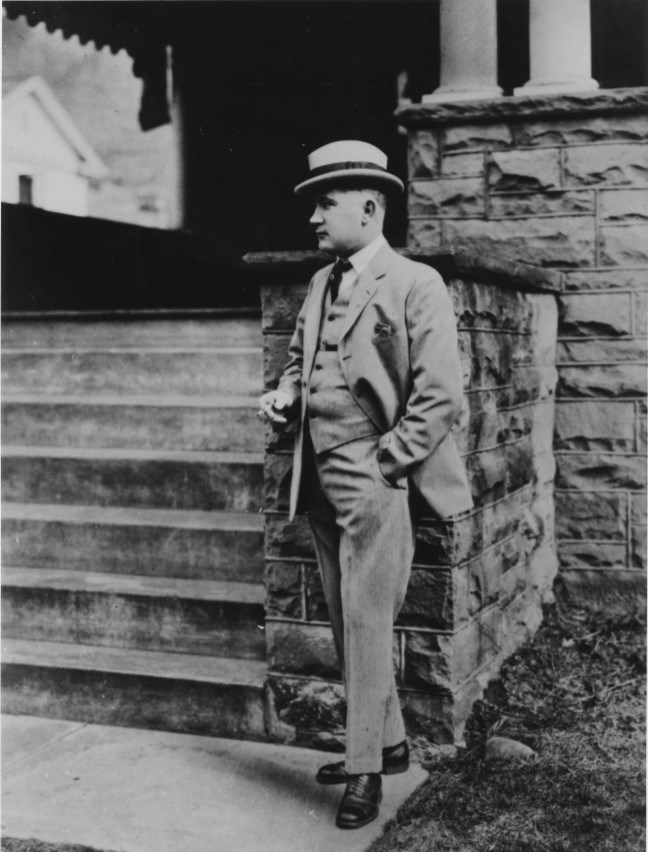 A man in a tan suit and hat leans on a stone pedestal near steps.