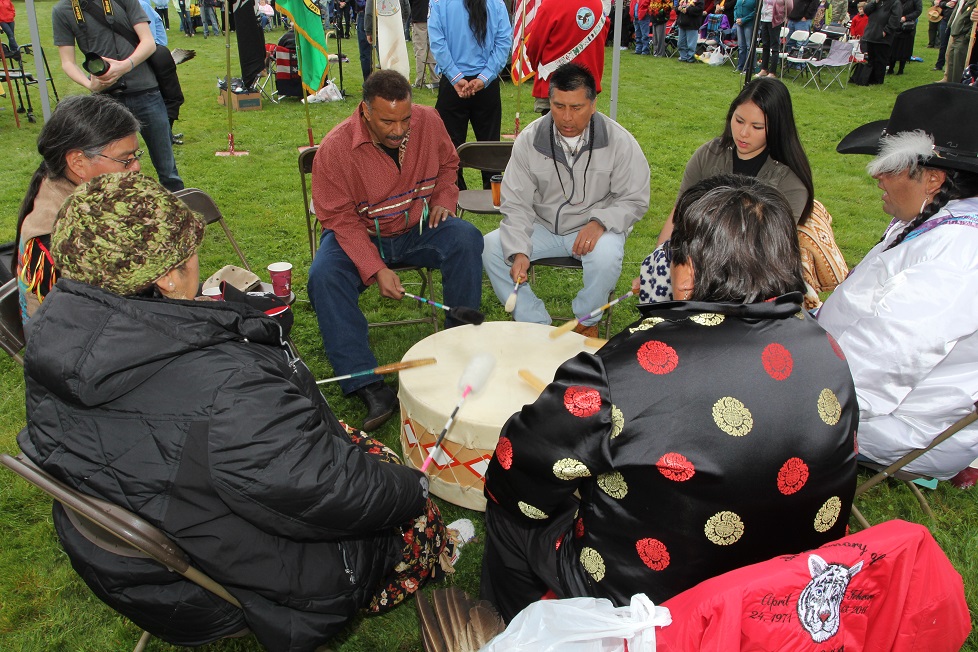 A group of men and women sit in a circle around a drum. Each holds a drumstick. They are outdoors in a grassy area.