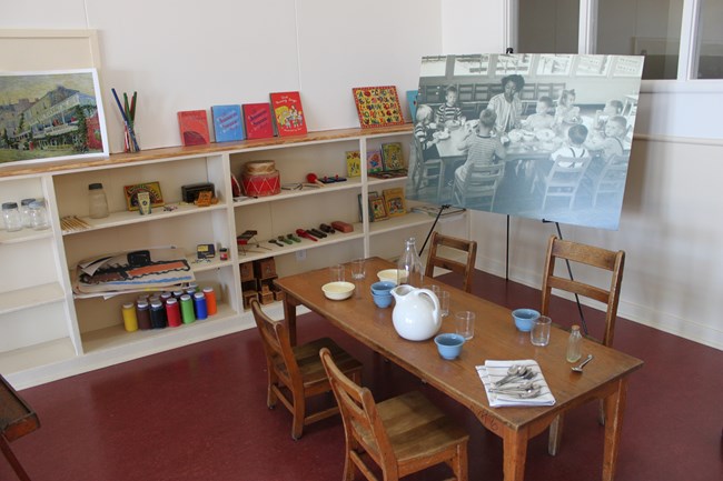 A small table with dishware sits in front of a shelf with toys and books. A blown-up photo of a woman sitting at a table with several children sits on an easel behind the table.
