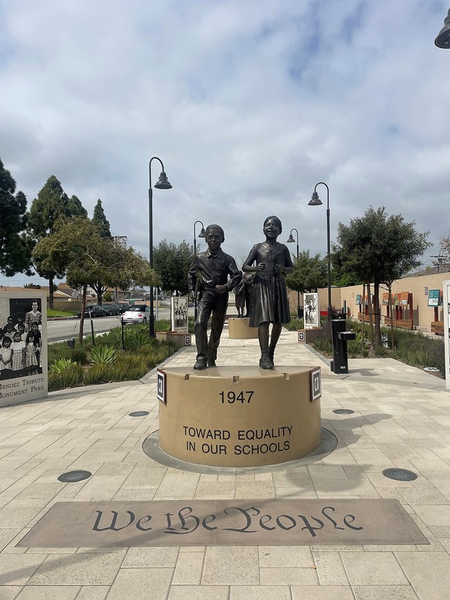 Statue of a young boy and girl walking with books in their hands. The base of the statue reads “1947: Toward Equality in Our Schools.” “We the People” is inscribed in script in the brickwork in front of the statue.