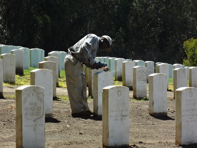 A man uses a wire brush on a power drill to alter the surface of a grave marker.