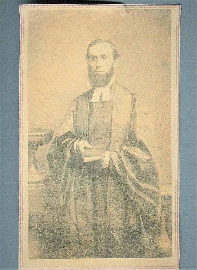 Photo of man, bearded, in black robes, holding a book