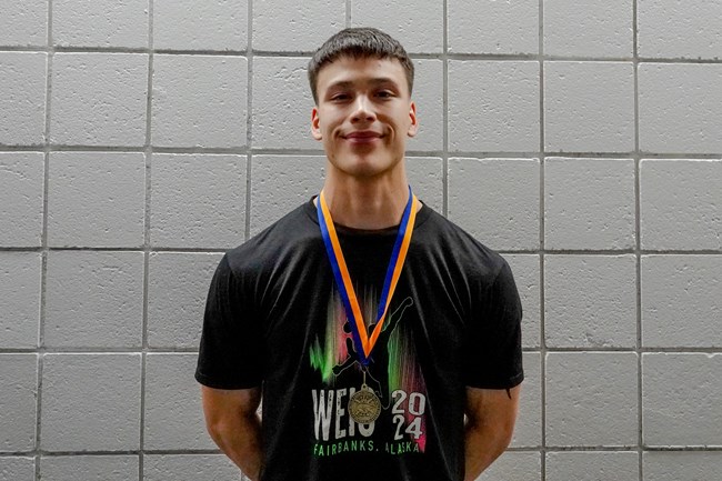 a young alaska native man smiles against the backdrop of cinder block walls.