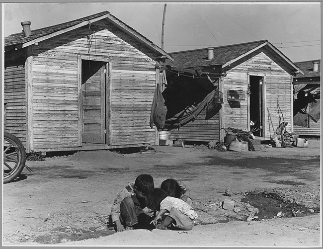 Black and white photo of three children playing in the dirt in front of small wooden cabins. Laundry is hanging from lines between the houses. Other items are strewn outside the entrance of another house.