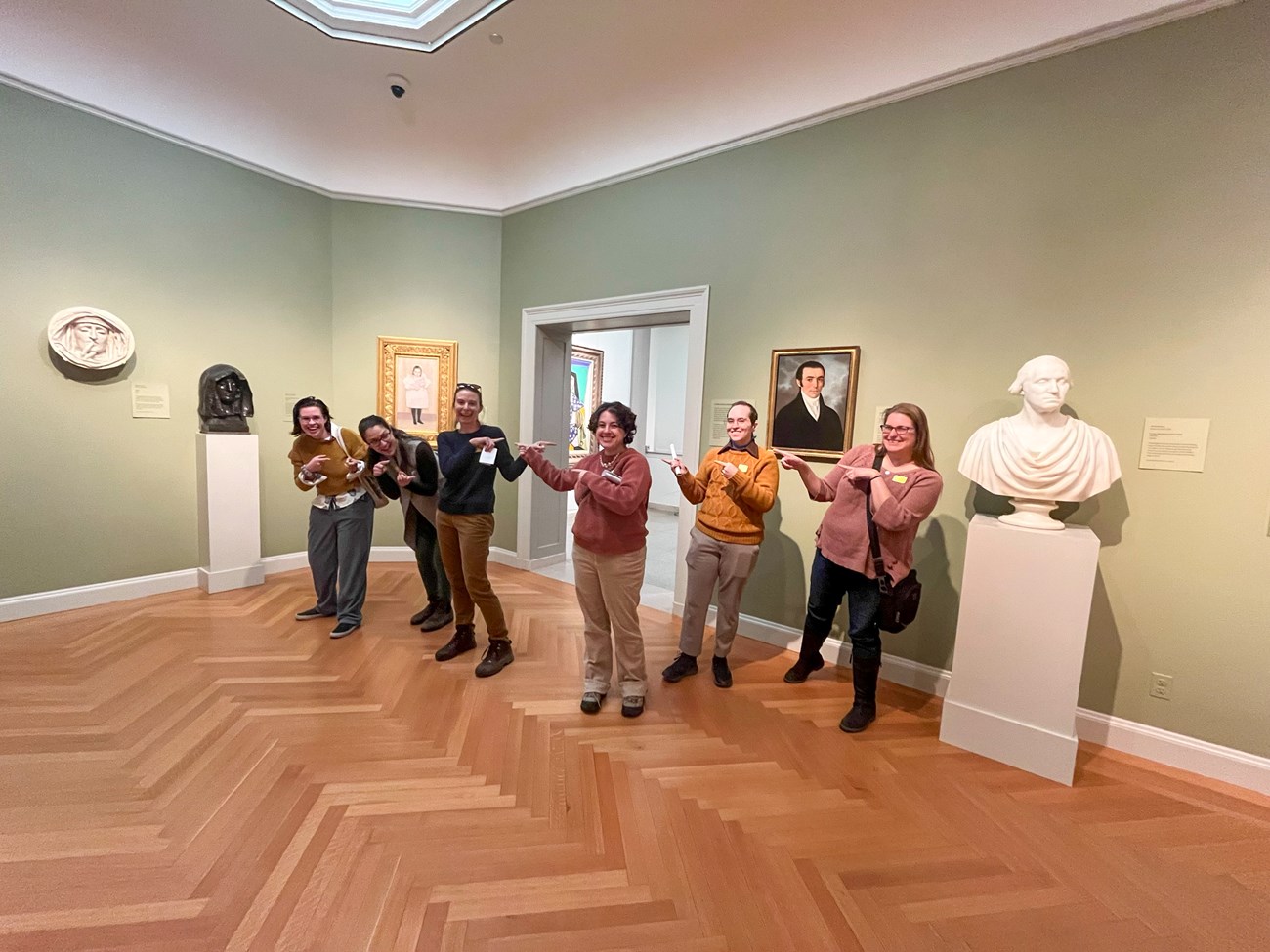 six people stand in art gallery museum exhibit pointing and each other and smiling