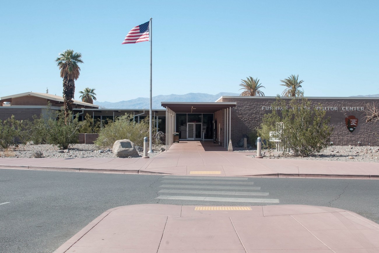 Furnace Creek Visitor Center front entrance. The pathway from the parking area to the entrance of the visitor center has curb cuts and tactile bumps. The visitor center is single level and mid-century modern design. The entryway has double doors.