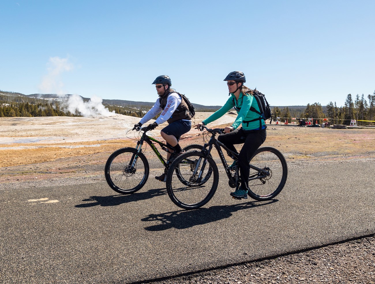 A man and a woman on bicycles on a paved road with a geyser erupting i nthe background