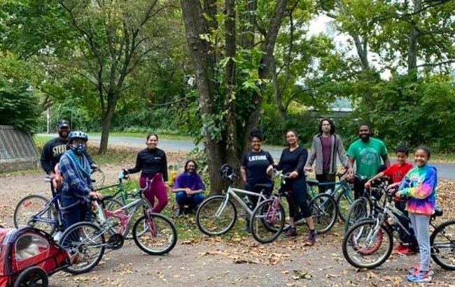Ten people of varied ages (adults and children) with bikes stand together in park environment with large tree behind