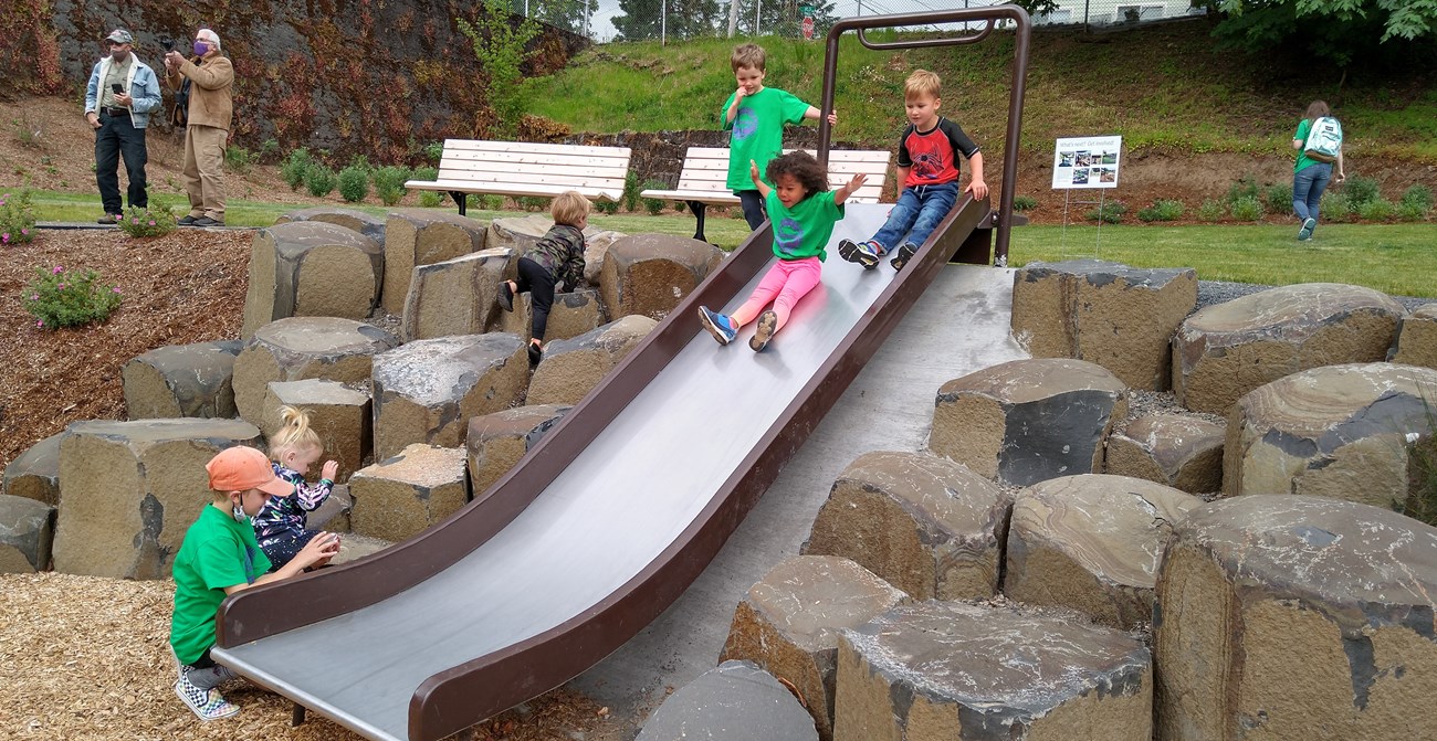 Small children play in D.C. Latourette park with a new slide surrounded by rocks and wood chips.
