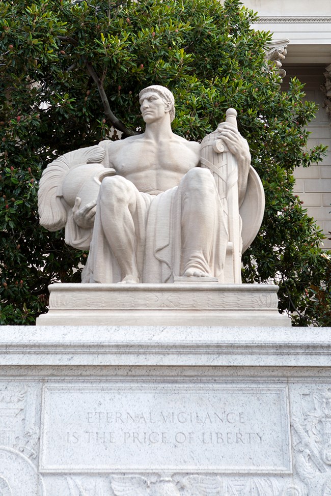 A statue depicting a seated man looking left with a serious expression on his face. He holds a sheathed sword at his right and a helmet at his left. At the base of the statue is, "ETERNAL VIGELENCE IS THE PRICE OF LIBERTY."