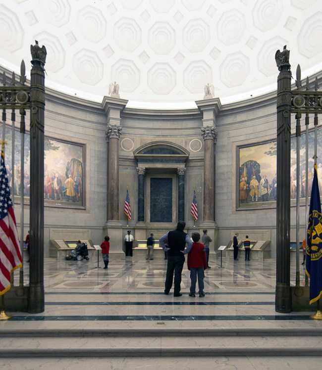 A grand rotunda with visitors in the background observing the Charters of Freedom. There are murals depicting scenes from American History on the walls. A boy and a man stand in the foreground observing the room.