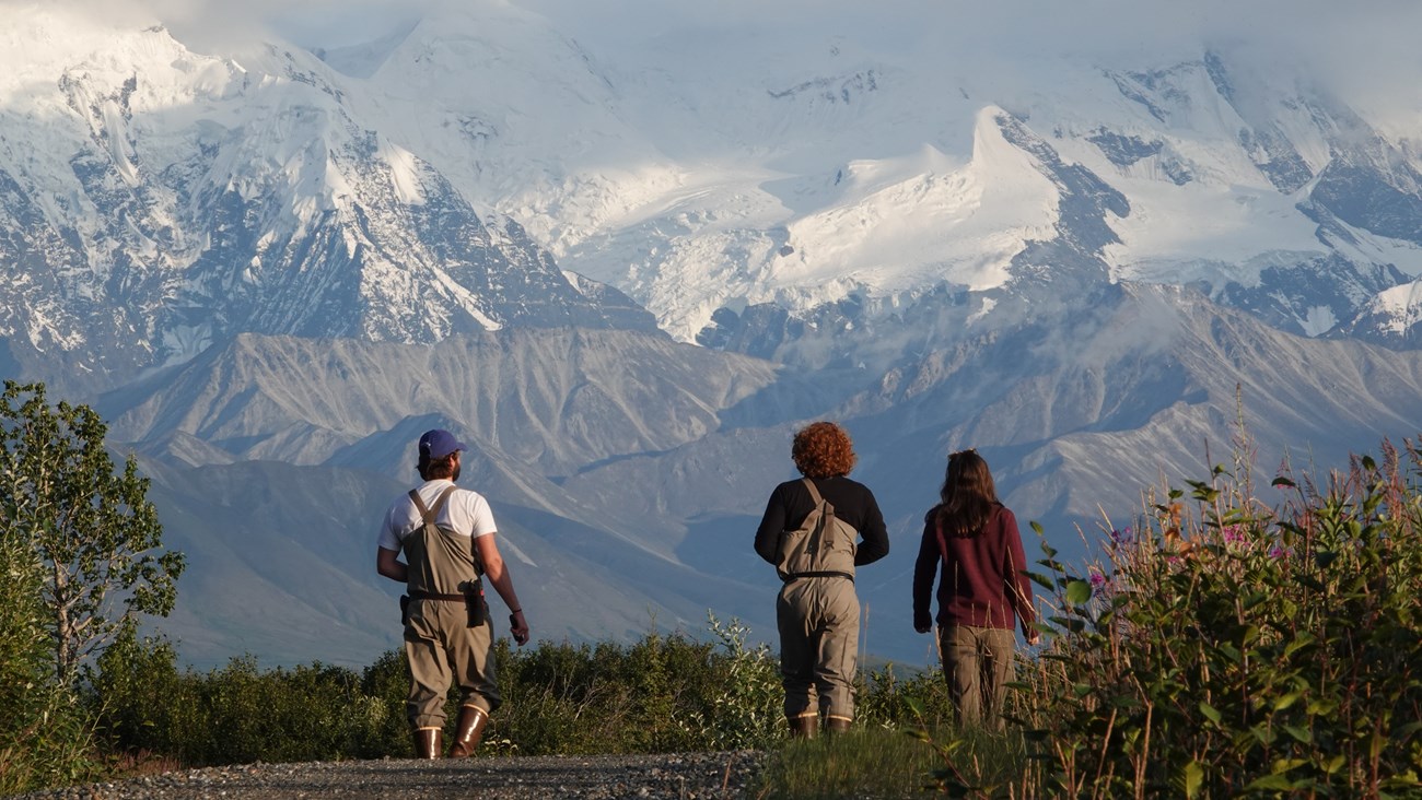 Young adults wearing waders face a mountain range.