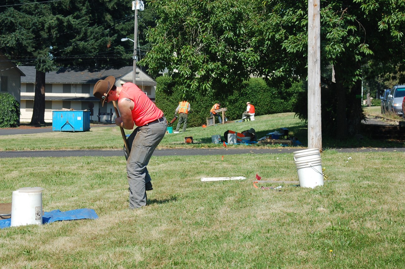 An archaeologist is shown digging a small hole with a shovel in the foreground. Three archaeologist are shown in the background. Two are moving dirt and screening for artifacts while one is sitting on an upturned bucket completing paperwork.