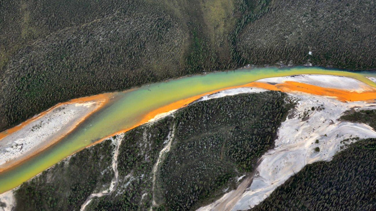 Aerial view of a braided river flowing from right to left. One braid entering the main channel is a bright, solid orange. Another is a yellow hue, where the orange has been diluted. A third is half yellow and half clear, dark water.