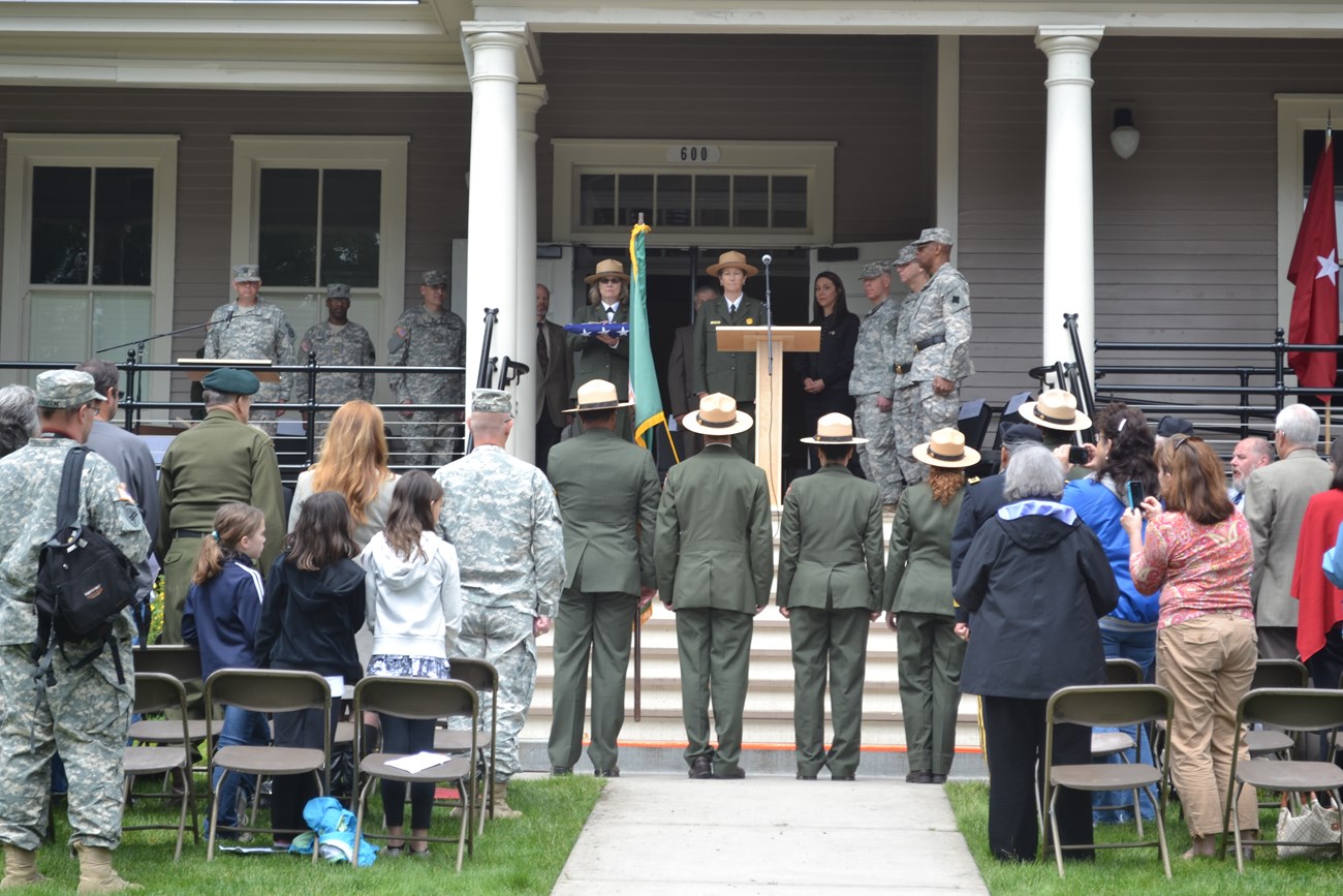 A row of National Park Service and US Army representative stand on the porch of a historic barracks building. One ranger is holding a folded flag. A line of National Park Service rangers stands at attention facing them.