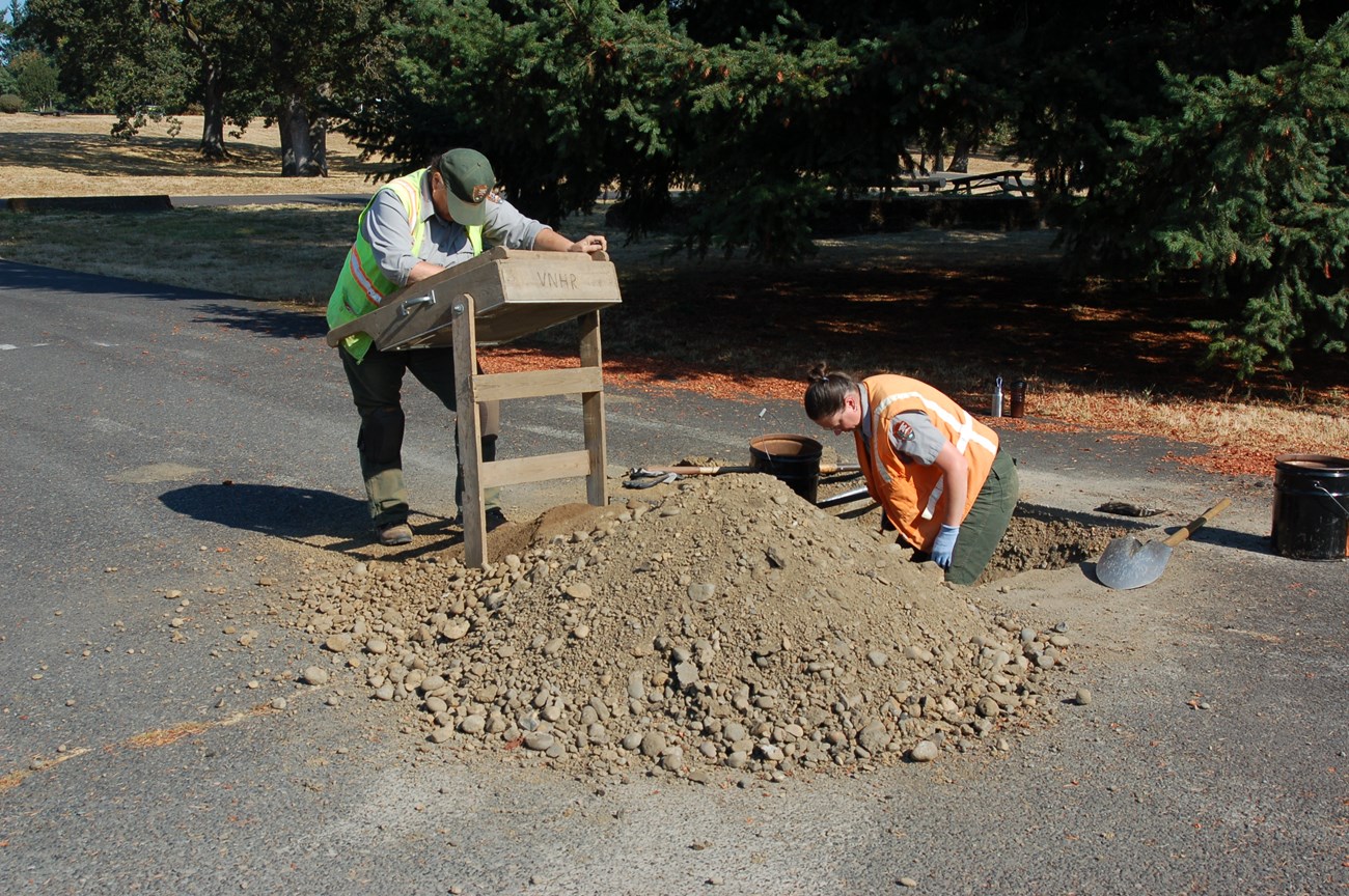 Two archaeologist are shown wearing National Park Service uniforms and safety vests. One is standing knee-deep in a square hole preparing to dig, and the other is leaning on an upright screen while looking for artifacts in the screened soil.