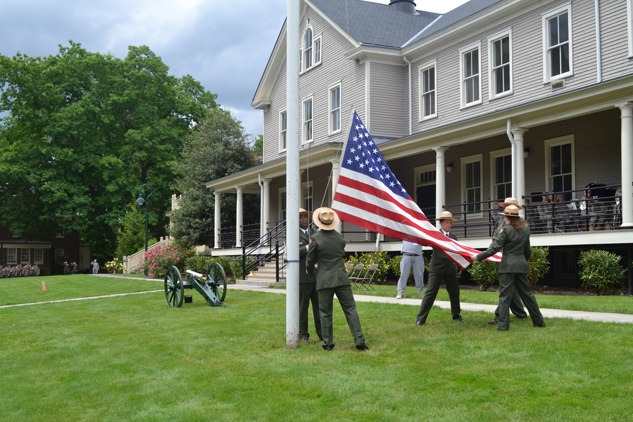 Five National Park Service rangers, in unform, raise an American flag. A historic barracks building and a cannon are in the background.]