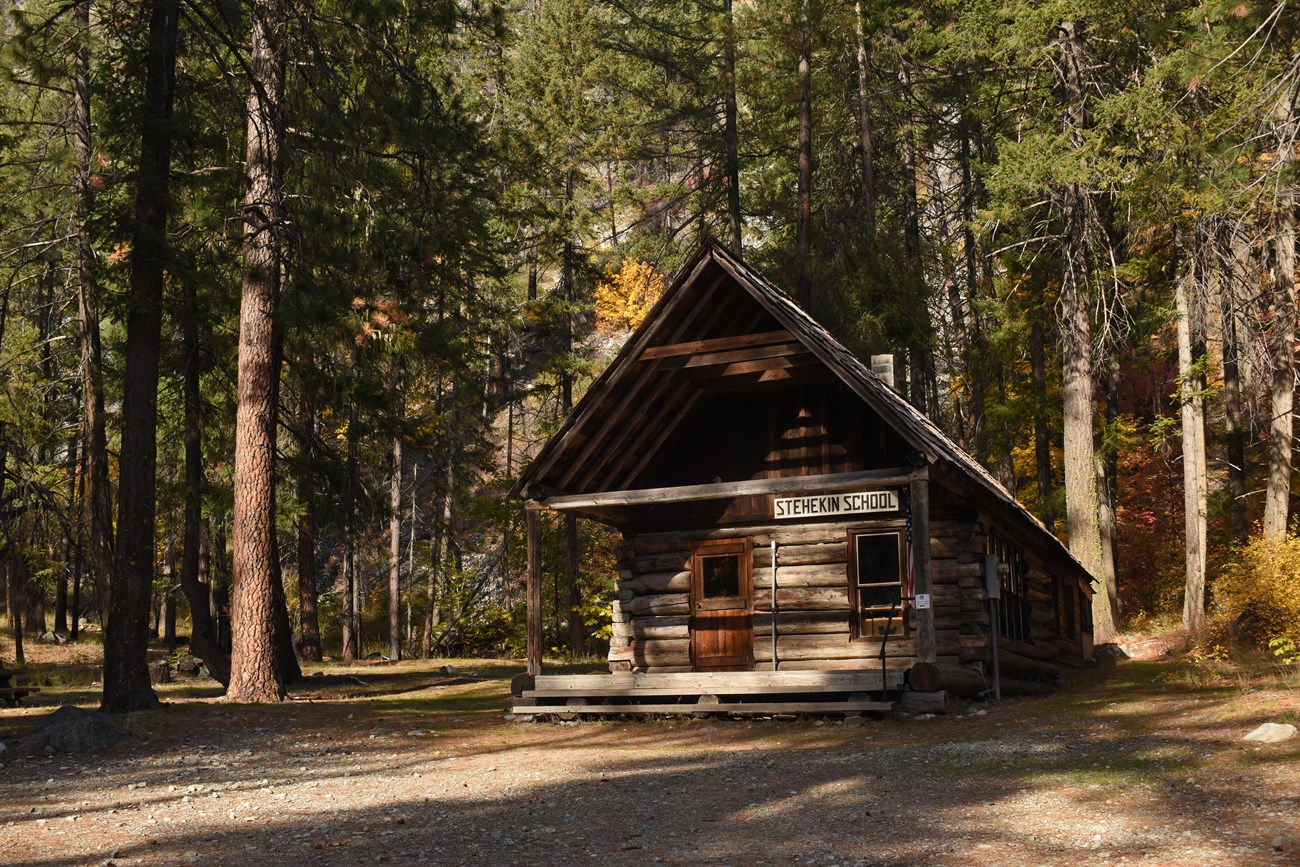 A wooden one-room school building with an American flag on a pole on the porch, sits in a sparse forest.