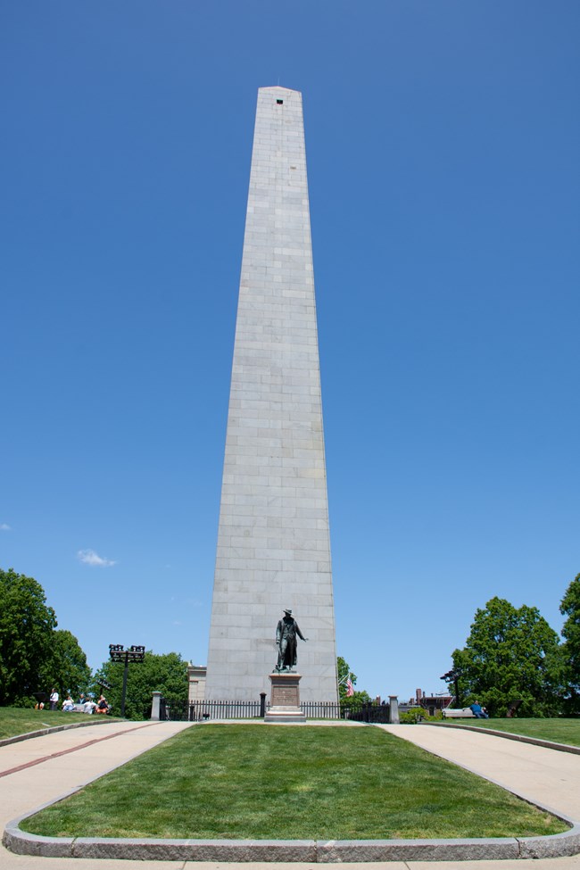 Bunker Hill Monument, a granite obelisk, standing before a bright blue sky. A statue of a man stands in front of the Monument.
