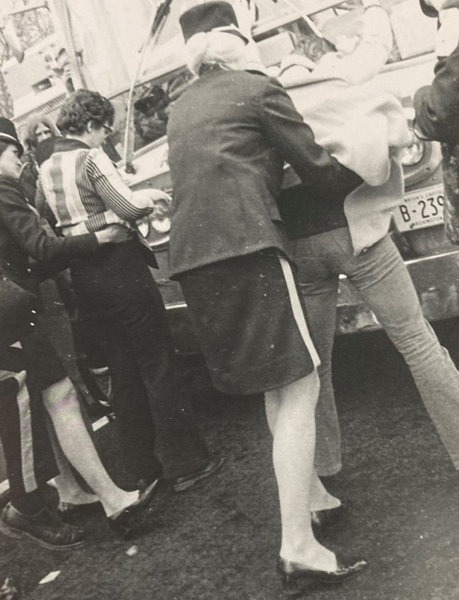 Woman in a police uniform with skirt pats down a woman demonstrator leaning against a bus.