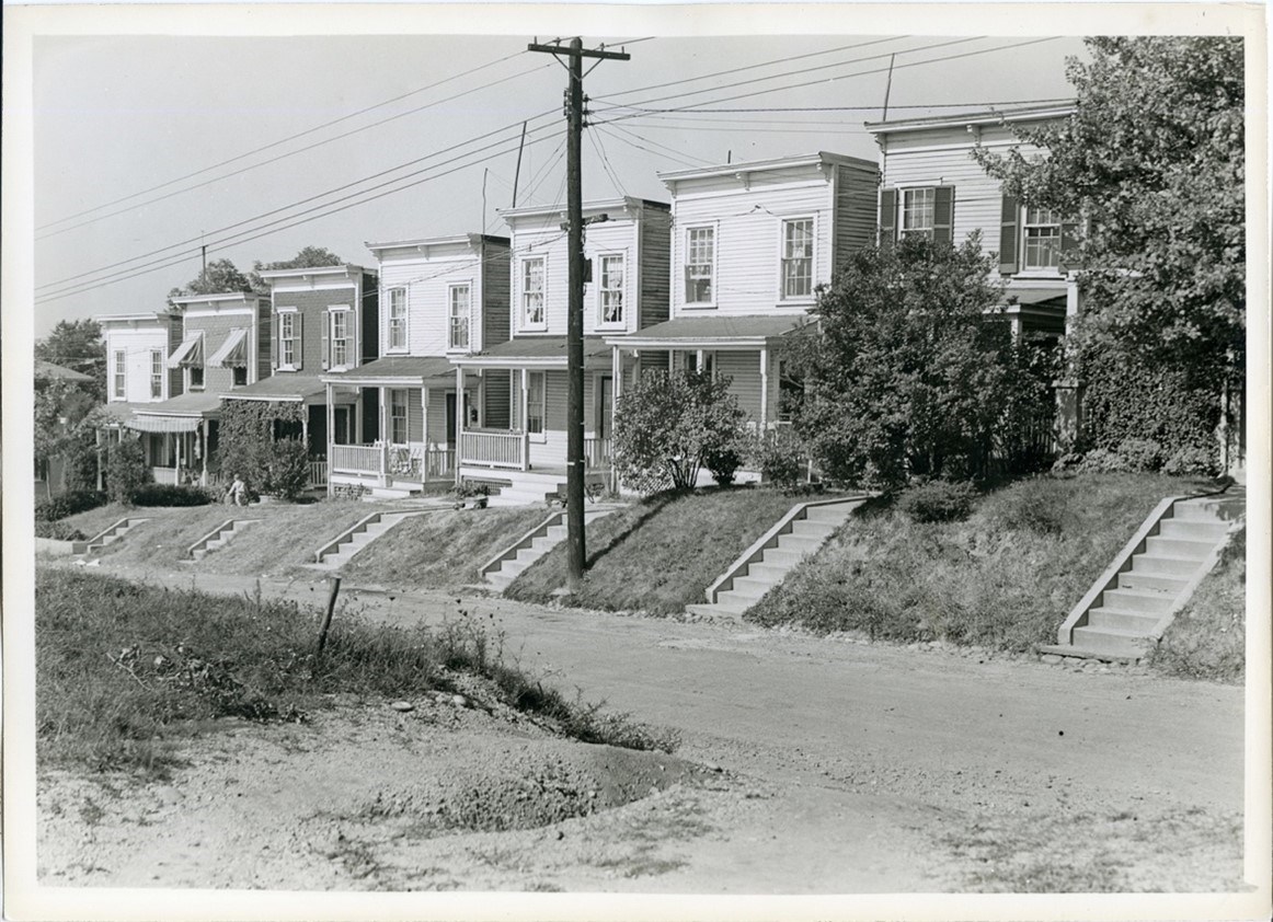 A row of seven houses with flat tops. stairs descend from each porch to a dirt road down a small grassy hill