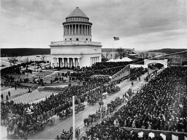 Black and white photo a birds eye view of a crowd of people seating in front of a large white mausoleum