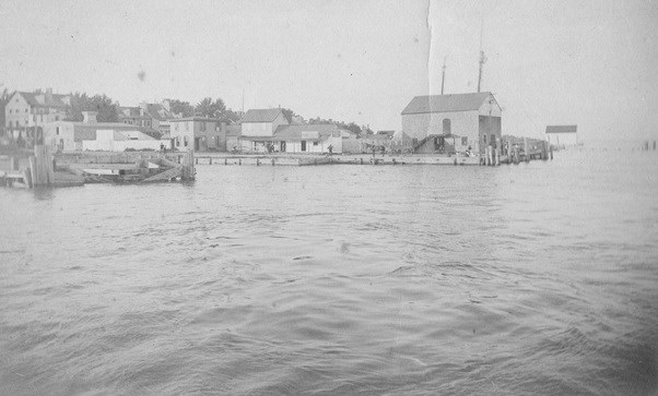 Black and white photograph of docks and a harbor with homes in the background.