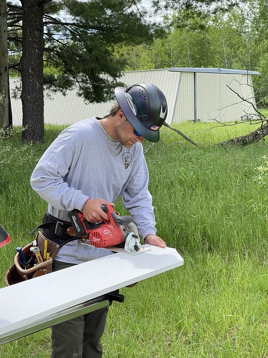 Worker wears a black hardhat with a brim, dark sunglasses and a grey national park service longsleeve shirt while looking down and cutting a plank of wood with a hand saw.