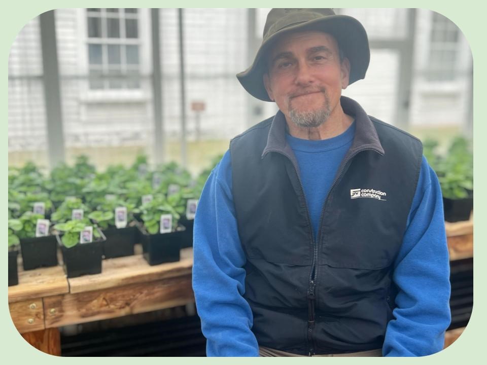 Chatham garden volunteer Mike Di Salvo seated inside one of the Chatham green houses with plant saplings in the background.