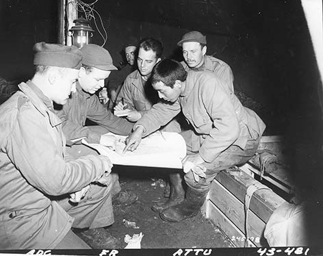 A Japanese man sitting down lean over a map is pointing to places on the map. There are 5 military men gathered around him, looking at what he is pointing. All are sitting down. They appear to be in a large tent.