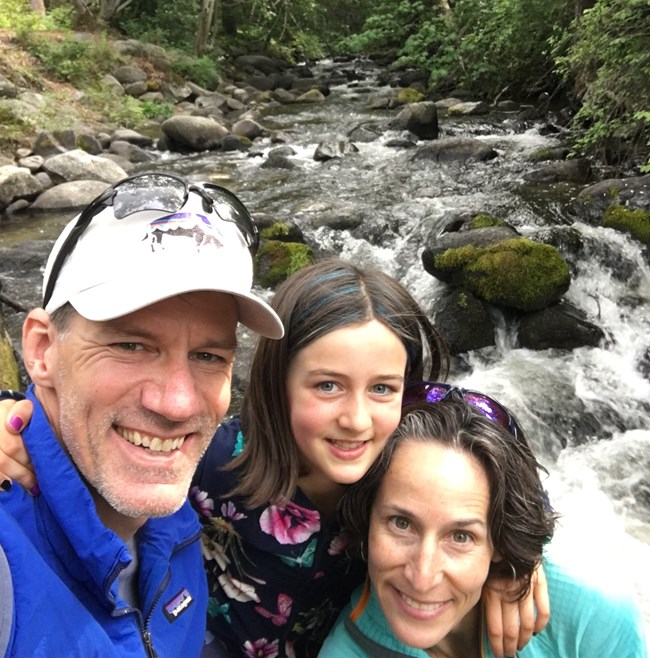 Man, woman and child hug each other in front of a stream.