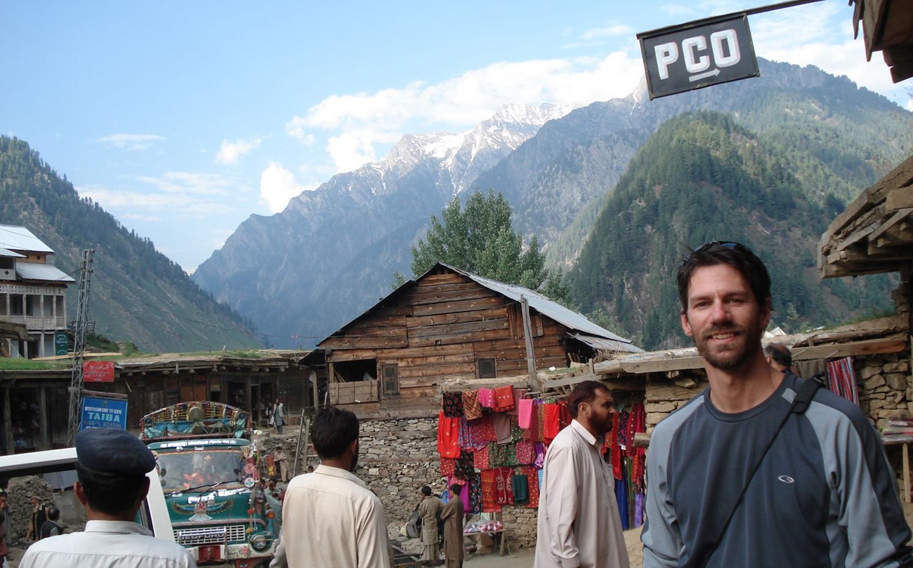 Smiling man on street of small village in Pakistan with colorful material hanging near a house and tall mountains in the background.