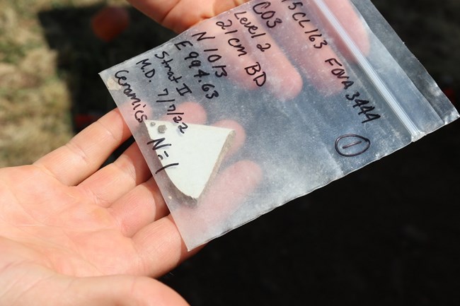A person holds a clear plastic bag with notes written on it in black ink. Inside the bag is a fragment of a white ceramic dish.