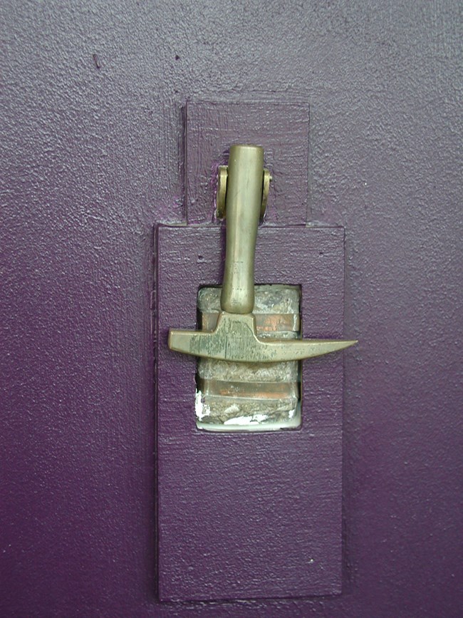 A door knocker in the shape of a rock hammer hangs on a purple door