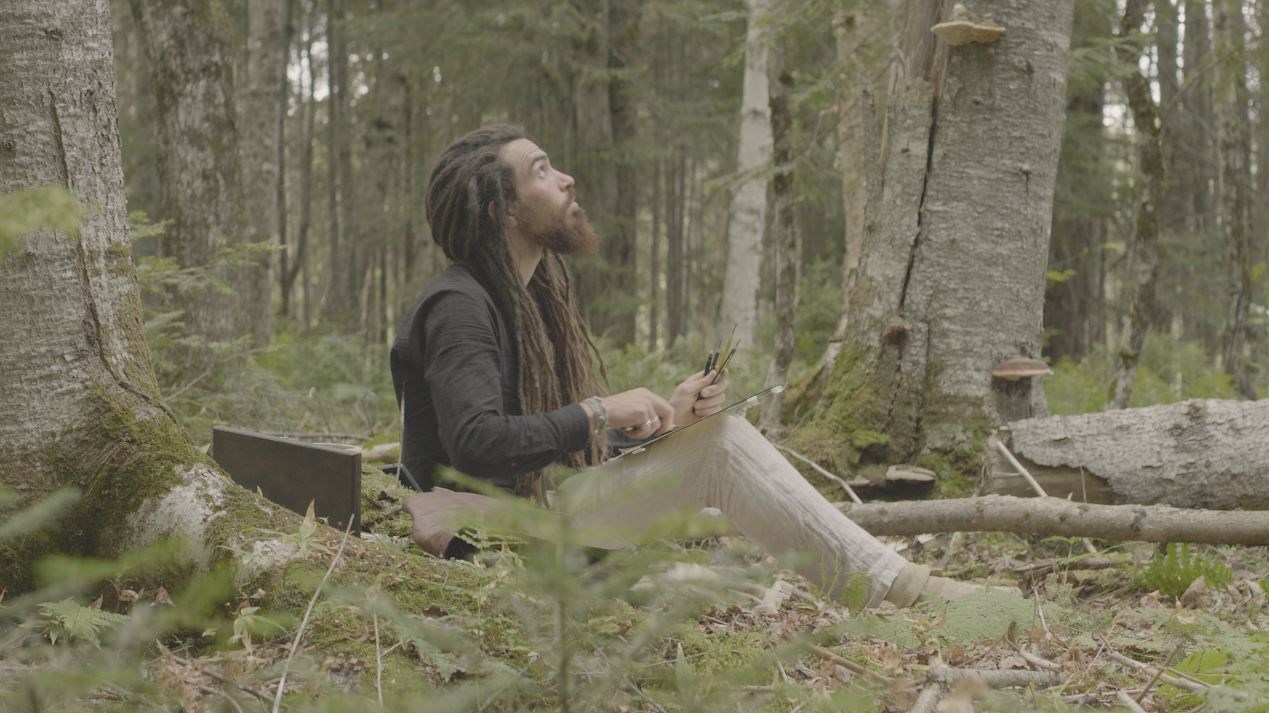 Man sitting on forest floor holding drawing tools looking up into canopy