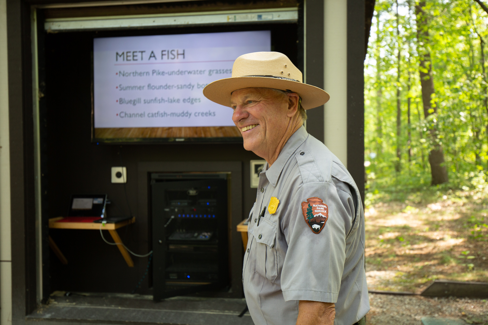 A ranger wearing a flat hat and a gray button-up shirt with an NPS patch smiles as he looks into the distance. Behind the ranger is a forest and a small building with a television screen that says "MEET A FISH".